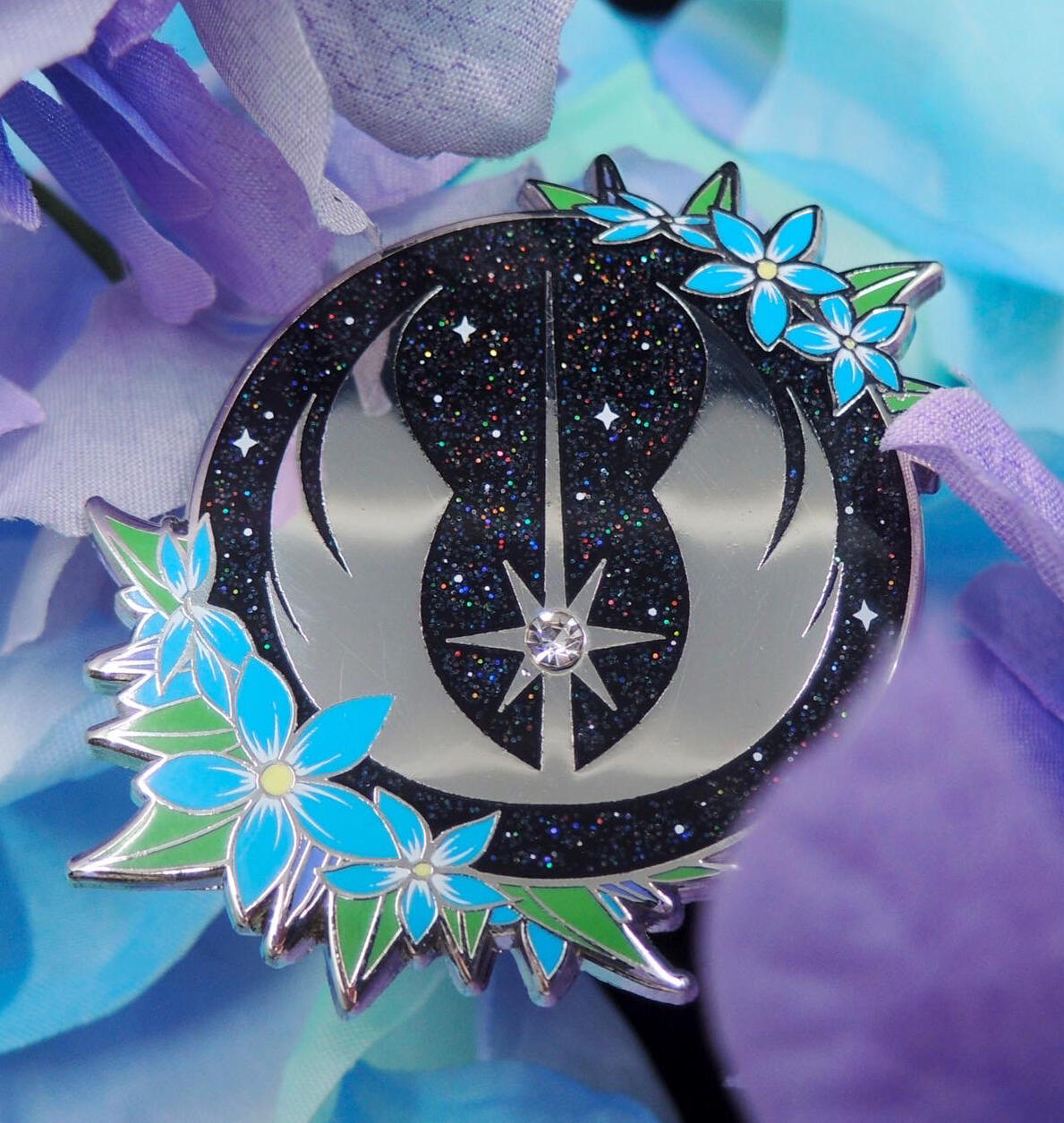 Photo of a round enamel pin, black with rainbow glitter and white printed stars. Around the outer edges of the circle are blue star-shaped flowers with yellow centers, purple buds, and green leaves. In the center is the Jedi symbol in silver metal, and it